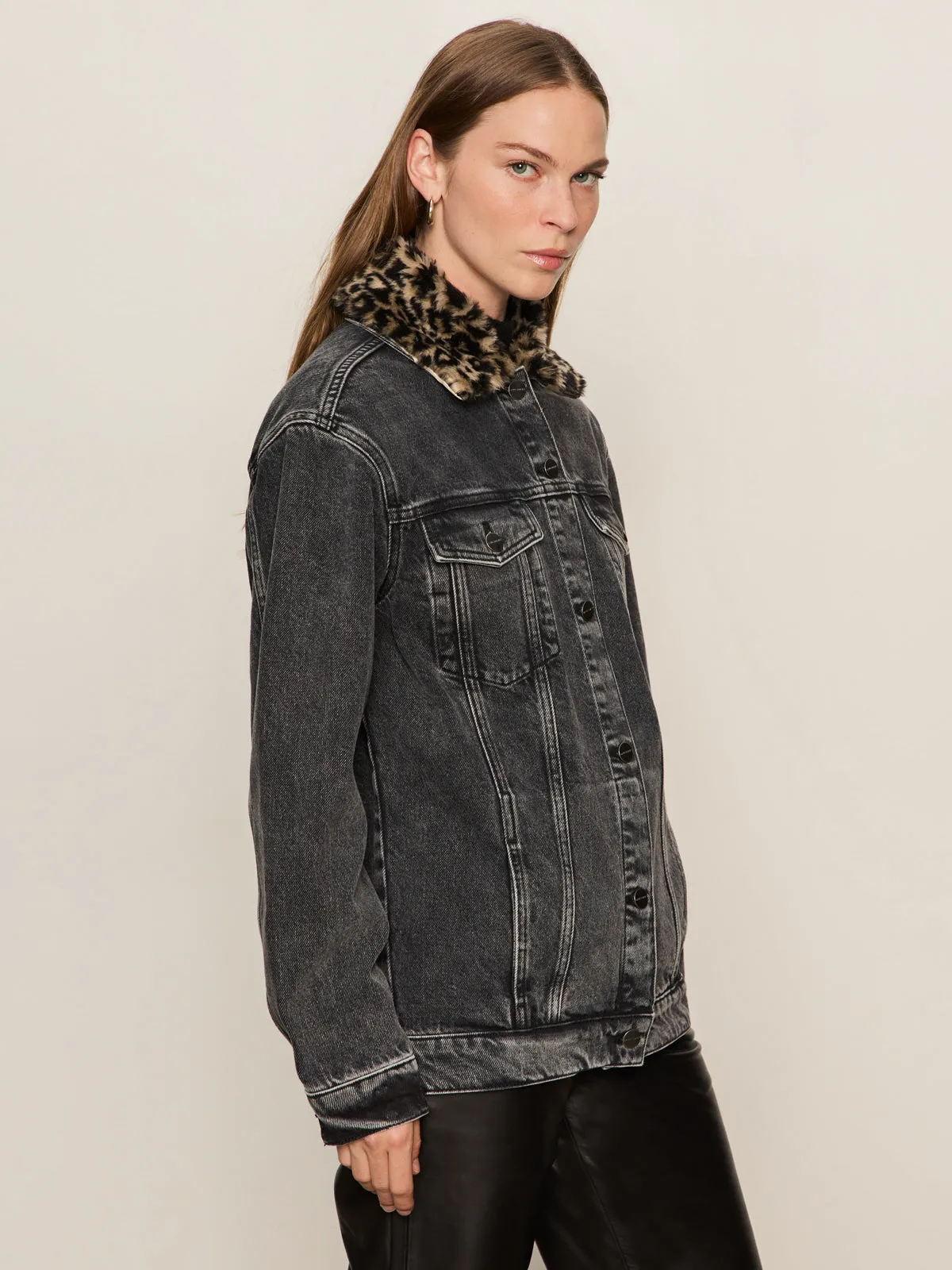 A person with long light brown hair wears the Sanctuary Clothing boyfriend trucker jacket in washed black denim, featuring a leopard print faux fur collar, paired with black pants, standing against a plain light background.
