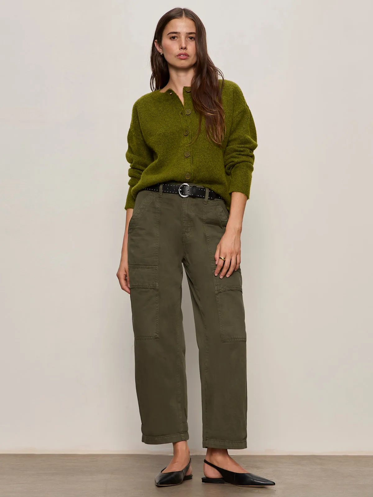 A woman with long brown hair and a neutral expression wears Sanctuary Clothing??s boyfriend utility pant in kalamata, styled with an olive sweater, black belt, and black pointed-toe flats against a plain background.