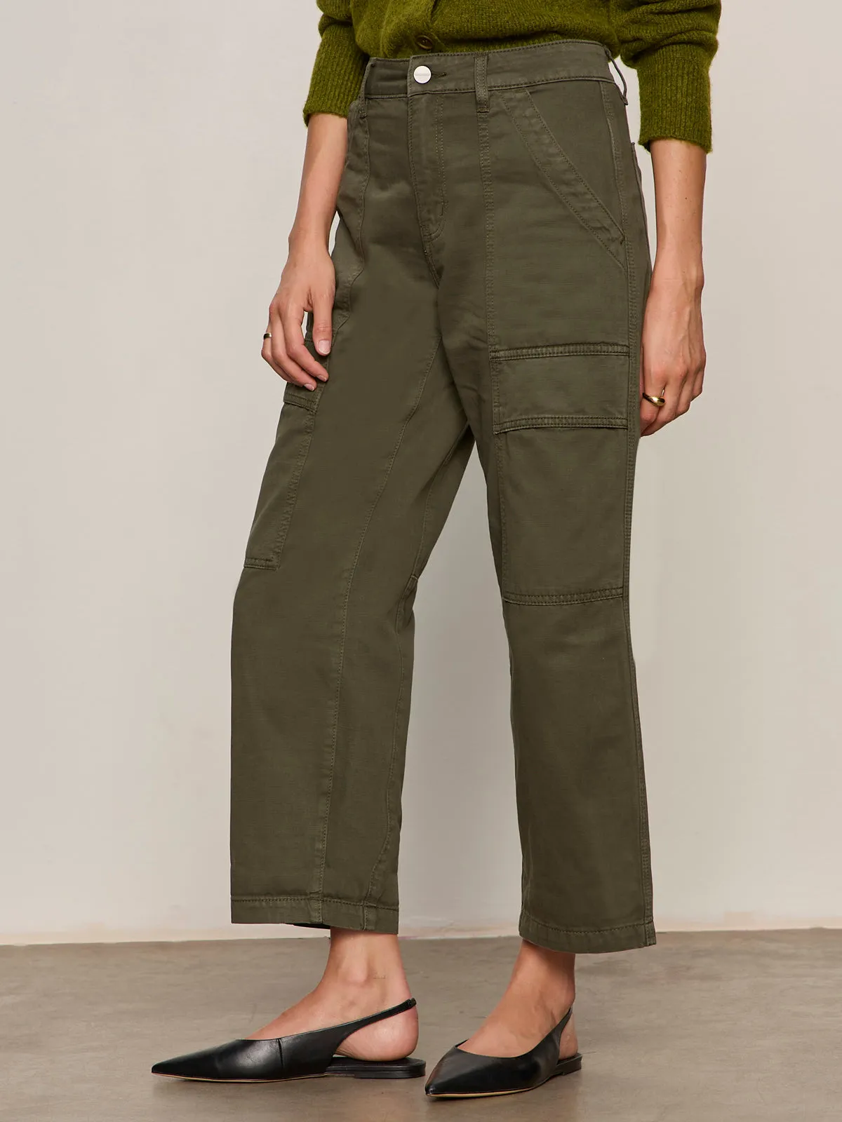 A person wears Sanctuary Clothing's boyfriend utility pant kalamata??olive green, high-waisted cargo pants with patch pockets??paired with a green sweater and black slingback flats, standing against a plain background.