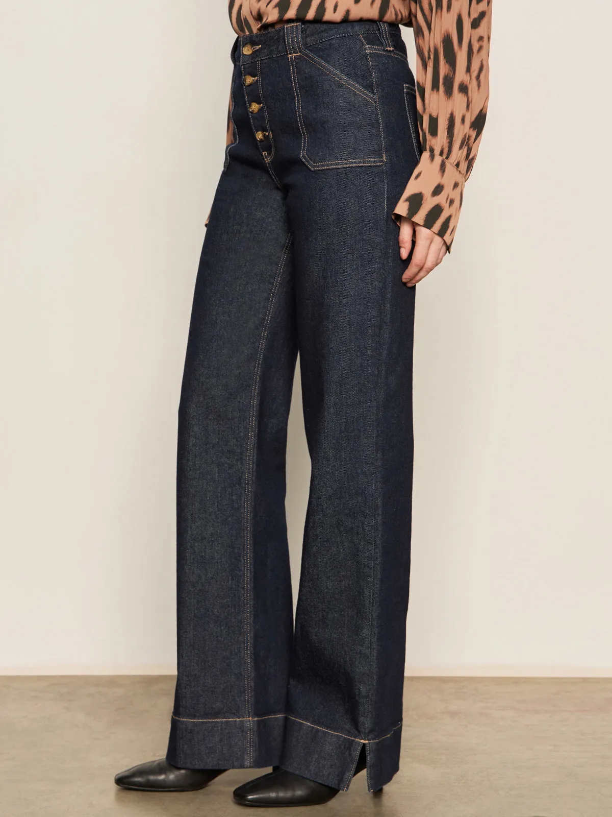 A person wears Sanctuary Clothing's ekara slim wide jean in indigo dark rinse wash, styled with a brown animal print blouse and black pointed-toe shoes against a plain neutral background.