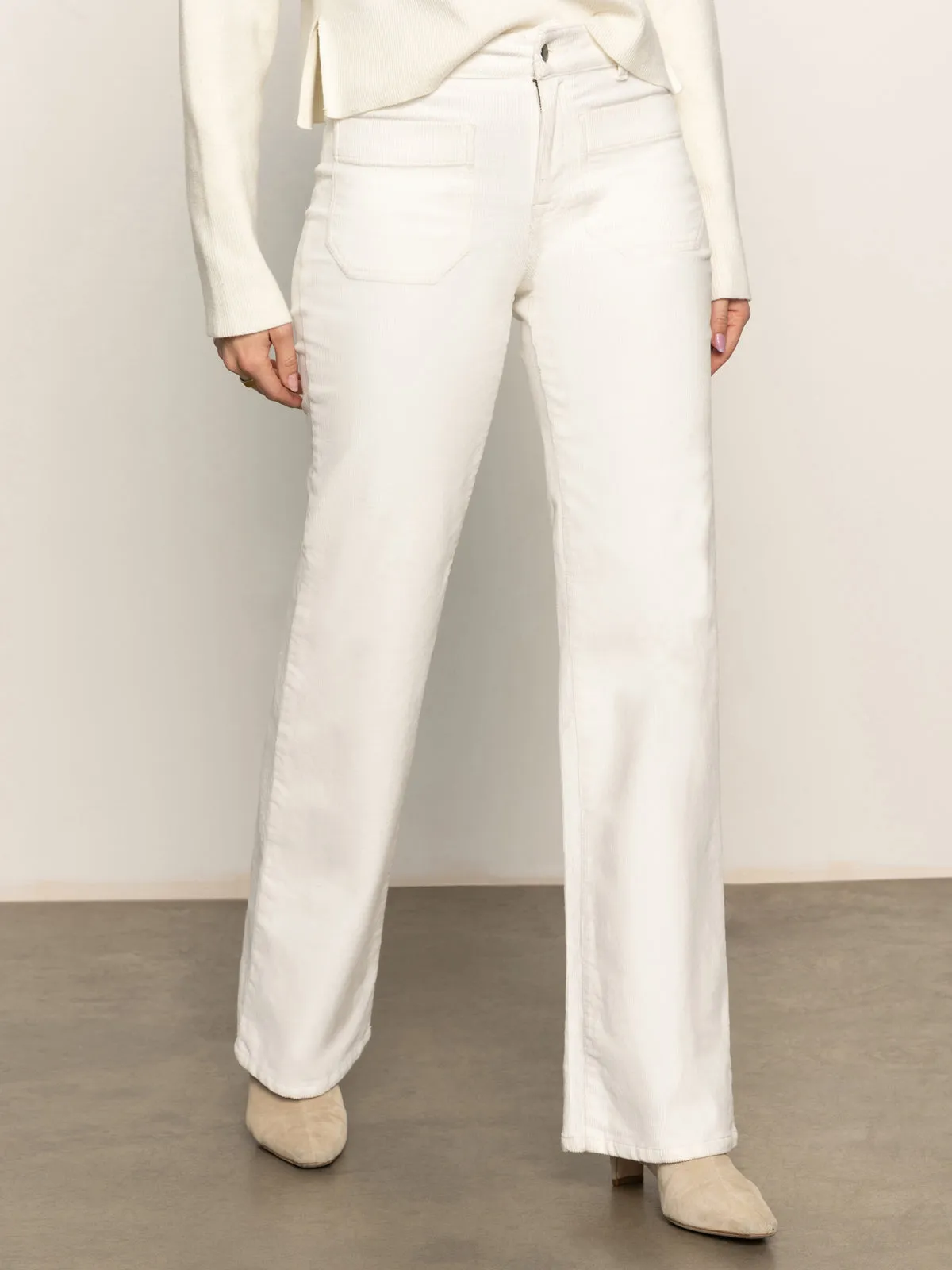 A person stands indoors against a neutral background wearing Sanctuary Clothing??s corduroy marine pant in chalk with a white long-sleeve top and beige ankle boots; their face is not visible.