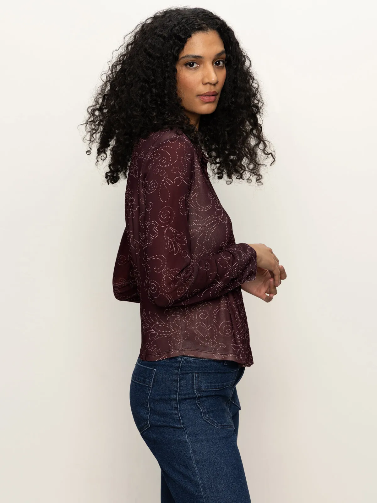 A woman with curly hair stands sideways, wearing the Sanctuary Clothing fitted mesh button down top boheme in maroon with blue jeans, against a plain light background.