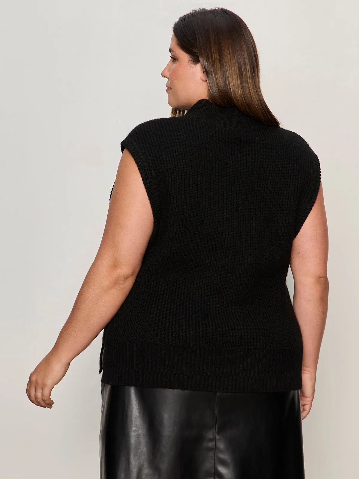 A woman with long brown hair is seen from the back wearing the Sanctuary Clothing city sweater shell in black (extended sizing) and a black leather skirt, standing against a light background.