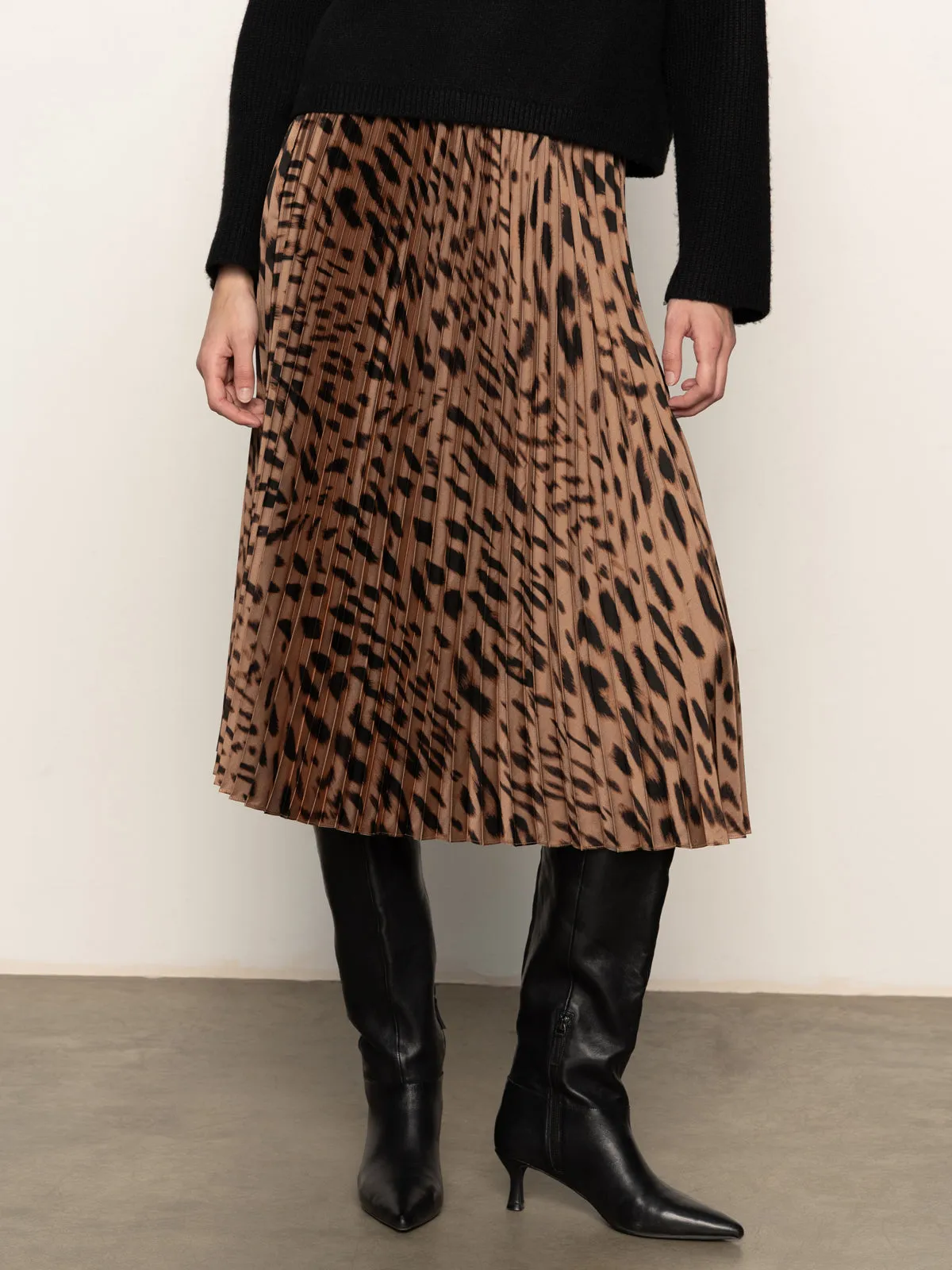 A person wears Sanctuary Clothing's nostalgia pleated midi skirt mocha leo, a black sweater, and black knee-high heeled boots, standing on a gray floor against a plain light wall.