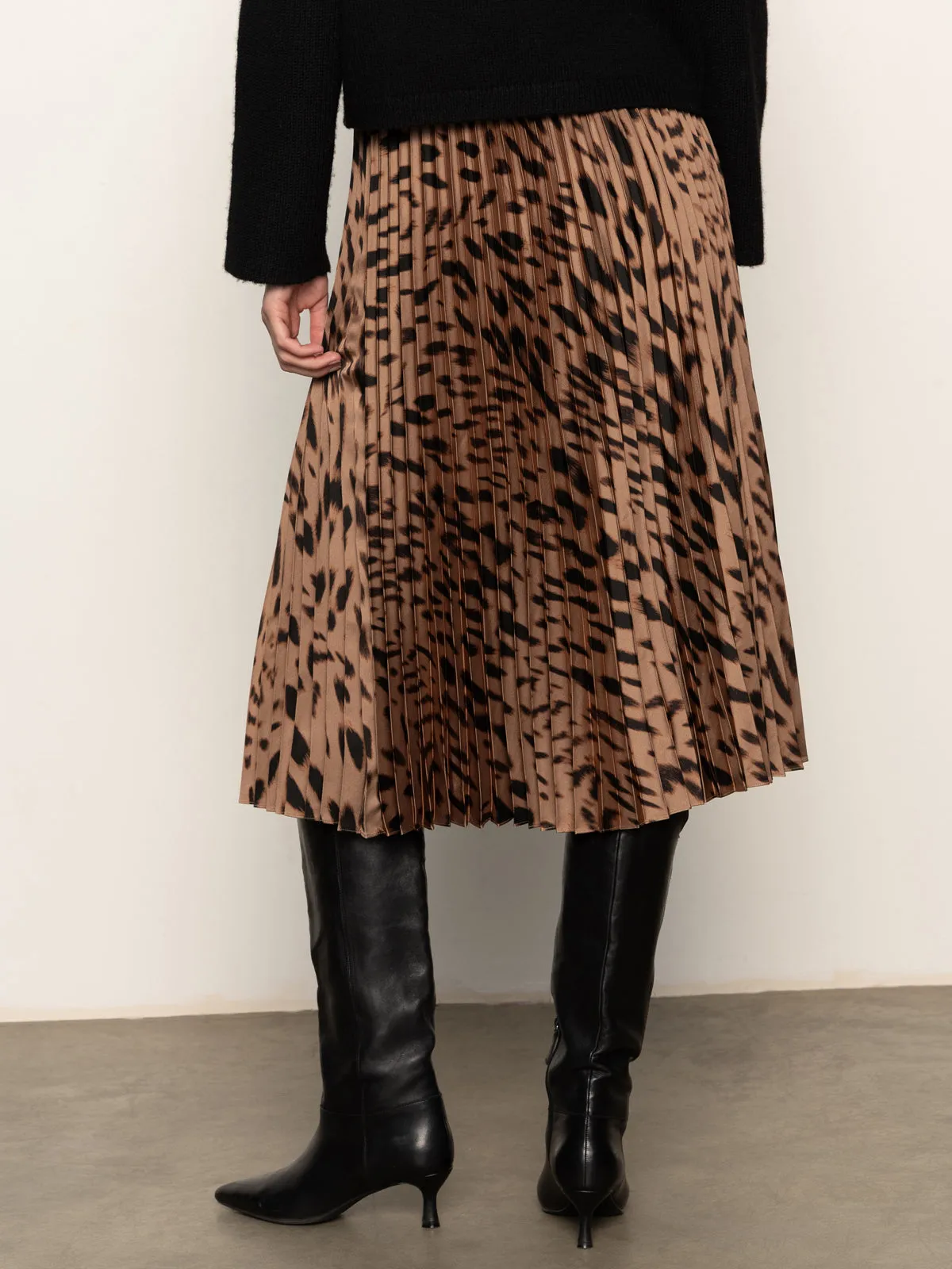 A person wearing the Sanctuary Clothing nostalgia pleated midi skirt in mocha leo, paired with knee-high black leather boots and a black top, stands on a gray floor against an off-white wall.