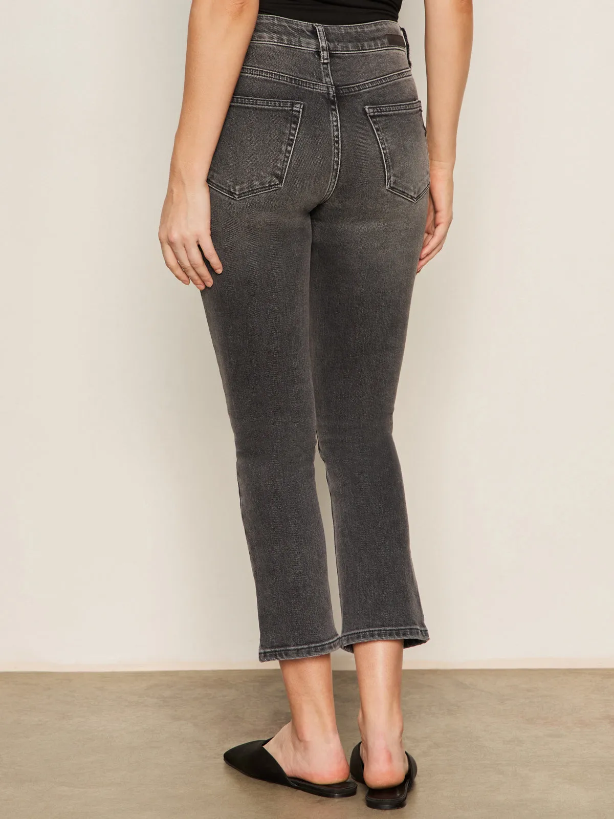 A person faces away, wearing Sanctuary Clothing's astra kick crop jean in washed black denim??high-waisted with raw hems and back pockets??paired with a black sleeveless top and slip-on shoes against a light, plain background.