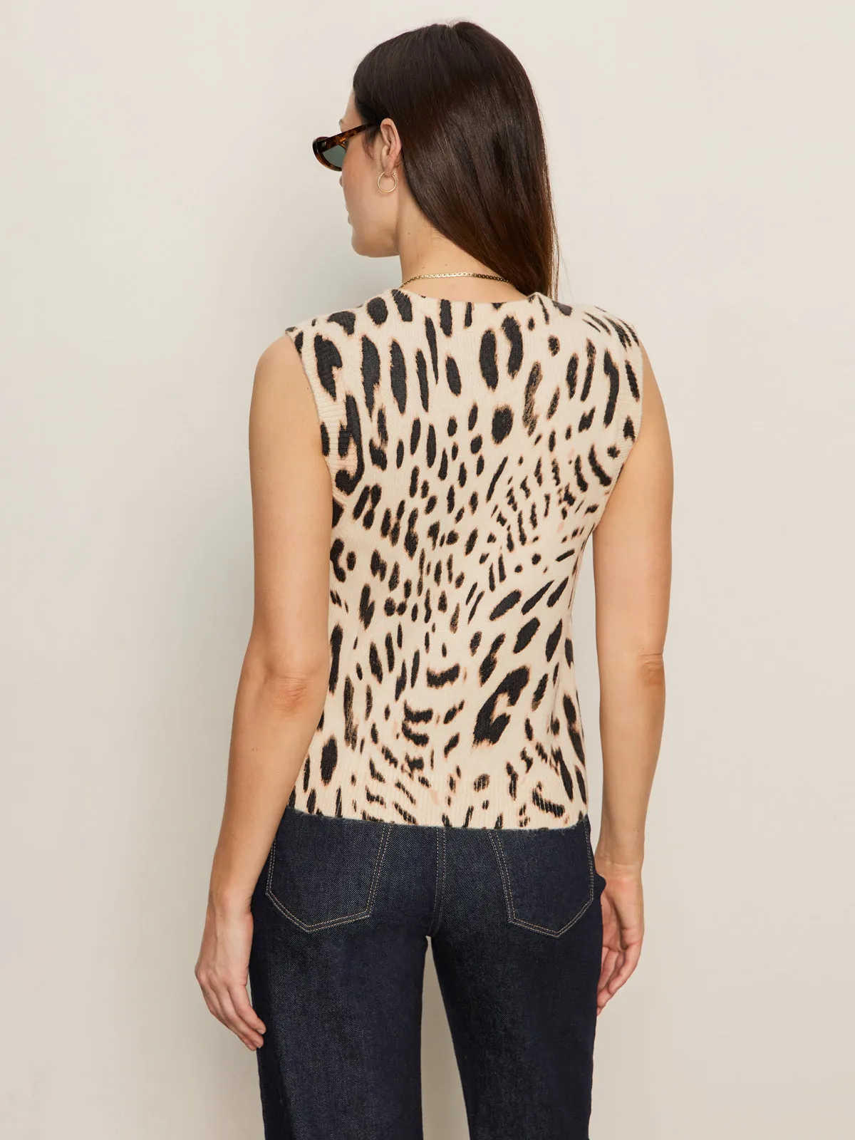 A woman with straight brown hair, wearing sunglasses, dark jeans, and the Sanctuary Clothing "fuzzy leopard shell mother leo" sleeveless top, stands facing away from the camera against a light-colored wall.