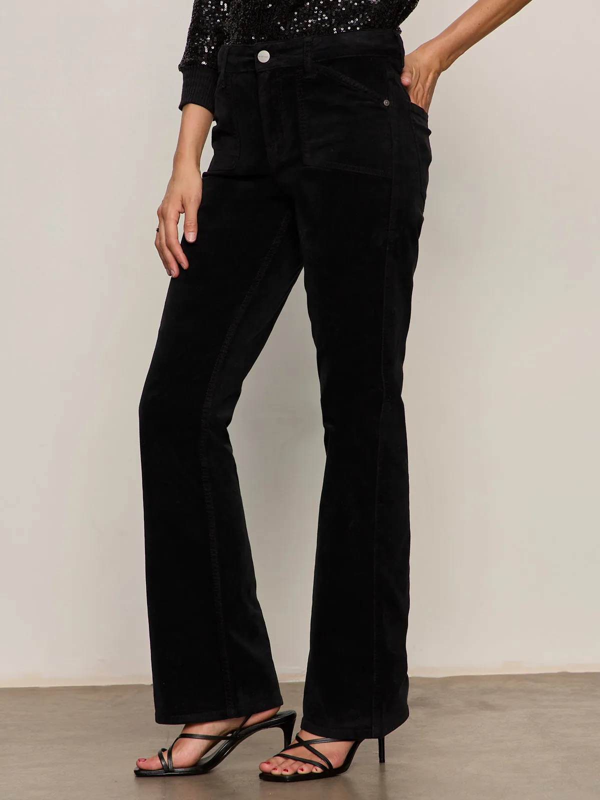 A person stands against a light background, wearing Sanctuary Clothing's velveteen brook bootcut pant in black petites, a black sequin top, and black strappy heeled sandals. Their left hand rests on their hip.